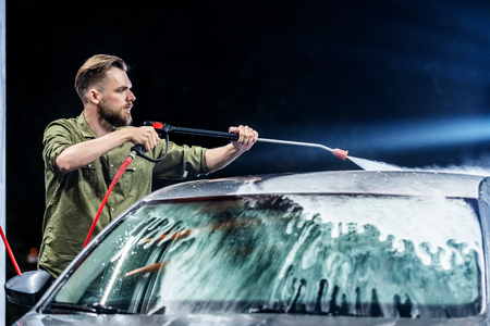A man with a beard washes a gray car with a high-pressure apparatus at night in a car wash. Expensive advertising photographyの写真素材