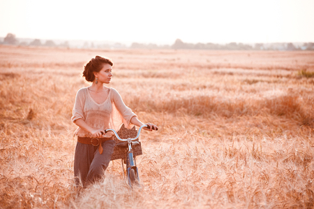 A beautiful girl in a golden rye field romantically comes with a bicycle on which gifts and looking on a free spaceの写真素材