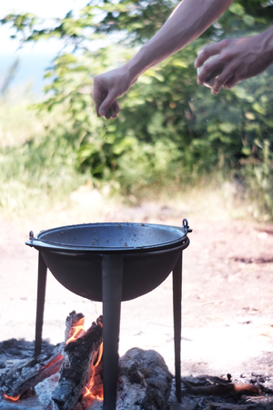 A man is cooking on fire. Hand pour salt or spices into the cauldron which stands on the fire with...