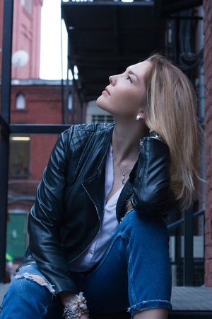 Young blonde woman with long hair is sitting on black stairs. Casual style leather jacket, jeans and blouse. Urban city dark backgroundの写真素材