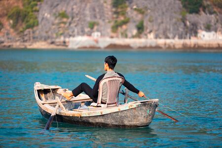Sole fisherman sitting on plastic chair and rowing with legs fishing in Lan Ha Bay near Cat Ba...