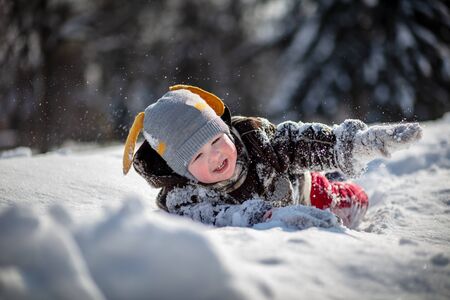 A little handsome boy in red pants and a brown sheepskin coat makes his way through the snowdrifts of snow. The child is being played. Trees in the snow in the background.の写真素材