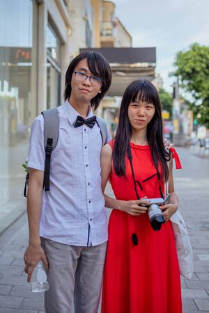 Tel Aviv. Israel. - May 2, 2017: Two Chinese tourists walk along the streets of Tel Aviv. A man with a backpack, in a blue shirt and pants. A woman in a red dress holds a camera in her hands.の写真素材