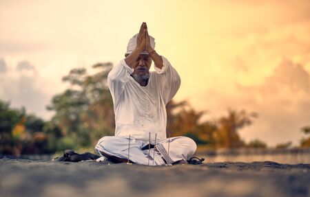 Bali, Indonesia - 2 Dec 2019: Old local balinese man in meditation near the sea. Concept of pray. Adult practice yoga on the Keramas black sand beach at sunset. Rural asian man doing yoga exercise on the beach at sunset.のeditorial素材