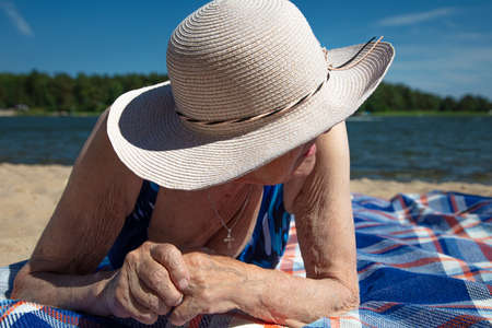 An elderly woman, in a summer hat on the beach. Model backs. Sunny, summer day by the sea. Concept:...