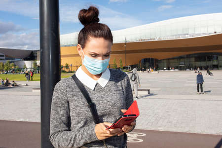 Woman in a medical mask, with a smartphone, brunette. Photo in the city. Summer day. High quality photoの写真素材