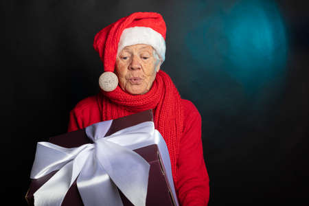 Surprised happy grandmother in a gnomes cap is holding a New Years gift. On a black background, studio photography. Congratulations on the new year 2021. High quality photoの写真素材