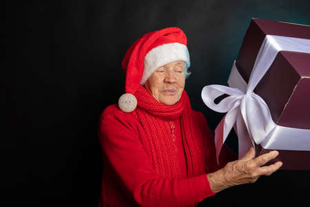 Sly, happy grandmother in a gnome cap is holding a New Years gift. On a black background photo studio. Happy New Year 2021. High quality photoの写真素材