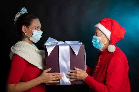 The girl in the outfit for the new year and grandmother in a gnome cap and medical mask are holding a New Years gift. On a black background photo studio. Happy New Year 2021. High quality photoの写真素材