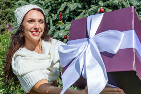 A young woman in a winter white hat near a decorated Christmas tree in the summer against a background of flowers with a huge gift. High quality photoの写真素材