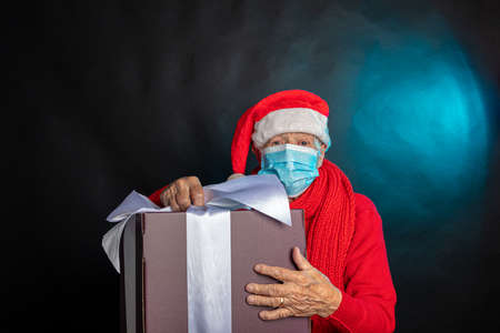 An old woman, in a medical mask, in a New Years cap, with a large gift in her hands. Studio photo. Dark background. Congratulations on Christmas. High quality photoの写真素材