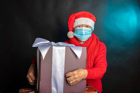 An old woman, in a medical mask, in a New Years cap, with a large gift in her hands. Studio photo. Dark background. Congratulations on Christmas. High quality photoの写真素材