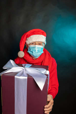 An old woman, in a medical mask, in a New Years cap, with a large gift in her hands. Studio photo. Dark background. Congratulations on Christmas. High quality photoの写真素材