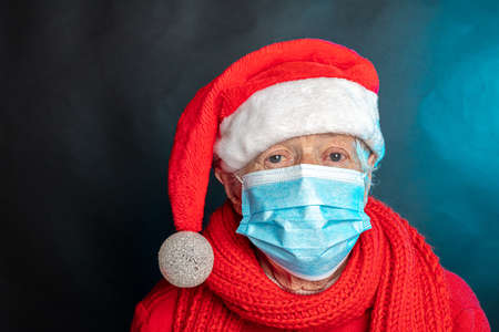 An old woman, in a medical mask, in a New Years cap, with a large gift in her hands. Studio photo. Dark background. Congratulations on Christmas. High quality photoの写真素材