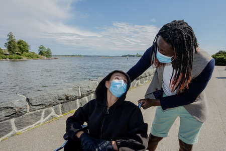 European women and African men wear a medical mask to protect themselves from the coronavirus outbreak. Woman in a wheelchair. Summer sunny day. Walk along the embankment. High quality photoの写真素材
