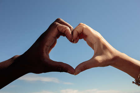 Close-up, fingers make a heart shape. Hand of a white and black man. Blue sky background. High quality photoの写真素材