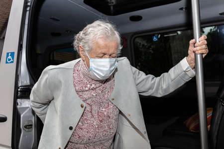 An old woman in a medical mask with a wheelchair, getting a disabled person into the car. High quality photoの写真素材