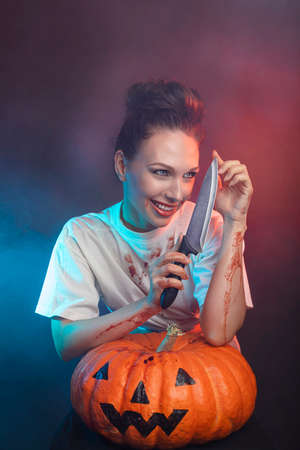 Woman covered in blood with a knife, dark hair and a pumpkin - halloween concept. High quality photoの写真素材