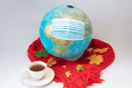The globe is wearing a medical mask and red warm scarf with maple leaves and a cup of tea. white...