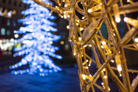 A street, Christmas garland is illuminated against the background of the New Year tree, where people...