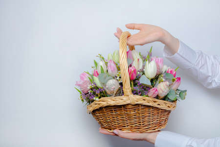 A woman in a white sweater holds a wicker basket with flowers on a white background, close-up without a face. Congratulations concept, there is a place for textの写真素材
