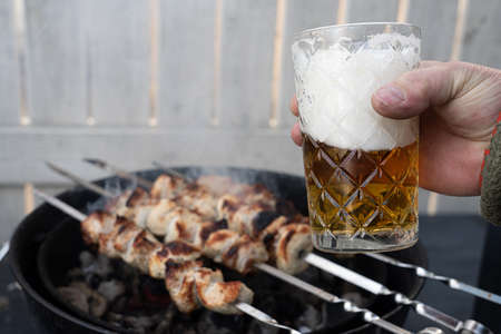 Glass of beer in hand on the background of a barbecue. Cooking blue pieces of meat on the grill. Traditional weekend picnic on a festive sunny day, harsh shadows. Street food.の写真素材