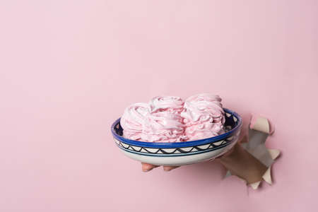 Hands hold a plate with homemade pink marshmallows through a torn hole on a pink paper background. Bakery advertising concept, recipe.の写真素材