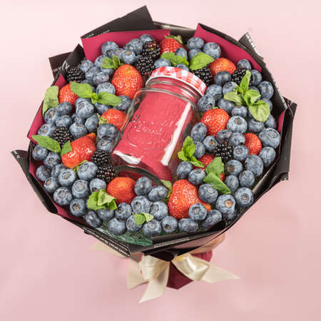 Hands hold edible berry bouquet with glass smoothie jar through torn hole on pink paper background. Concept for advertising, congratulations, recipe.の写真素材