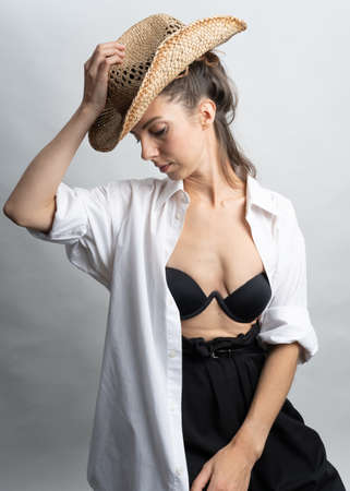 Model, brunette with a straw hat posing on a white background in the studio. She is wearing a white shirt, a black hairstyle.の写真素材