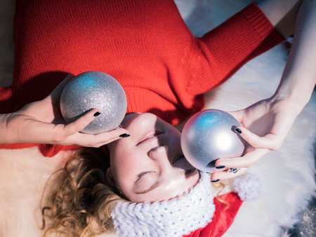 Portrait of a young woman with blond hair in a Christmas hat. She smiles. Close-up, lies on her back. Silver balls in the handsの写真素材