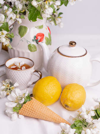 White tea set with tea in cups with yellow lemons and a blossoming apple branch. There are waffle cones on a saucer.の写真素材