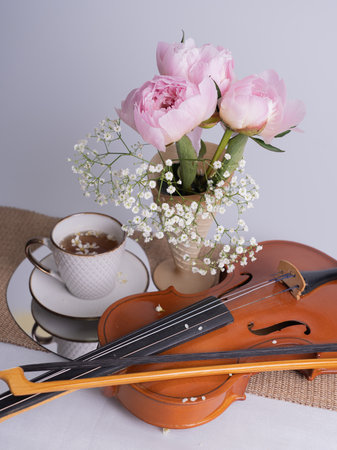 An artistic still life with a bouquet of pink peonies, a cup of tea and a violin. vertical photo.の写真素材