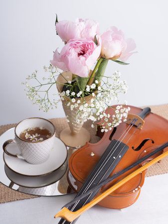An artistic still life with a bouquet of pink peonies, a cup of tea and a violin. vertical photo.の写真素材