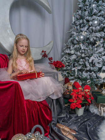 A little blonde girl in a pink dress opens gifts in a festive atmosphere against the background of a decorative moon. Magical Christmas time.の写真素材