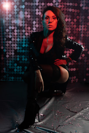A gorgeous brunette in a black bodysuit, black stockings and a sequined jacket at a party. nightclub. Red backlight in the dark. Photo with colored light.の写真素材