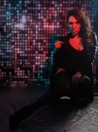 A gorgeous brunette in a black bodysuit, black stockings and a sequined jacket at a party. nightclub. Red backlight in the dark. Photo with colored light.の写真素材