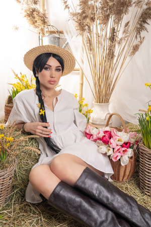 A beautiful brunette, with a long braid, she is in a white dress and a straw hat. Photo zone in the studio, On hay and daffodil flowersの写真素材