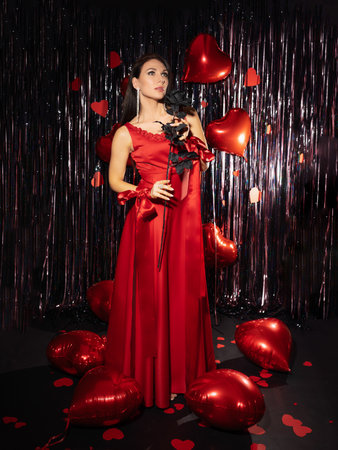 Beautiful, slender woman photo without a face in a red long, festive dress against the background of red balloons in the form of heartsの写真素材