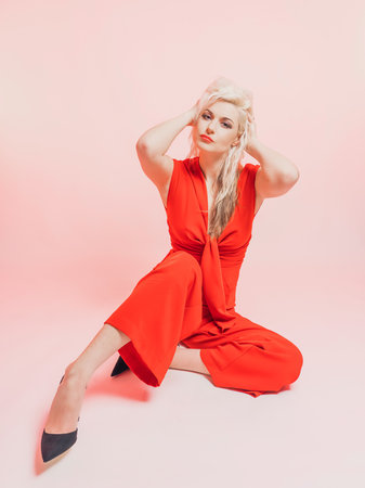 Portrait of a beautiful blonde woman in a red pantsuit. Posing while sitting in the studio on a pink background.の写真素材