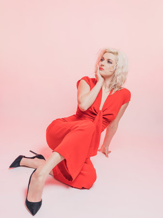 Portrait of a beautiful blonde woman in a red pantsuit.. Posing while sitting in the studio on a pink background.の写真素材