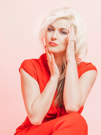 Portrait of a beautiful blonde woman in a red pantsuit.. Posing while sitting in the studio on a pink background.の写真素材