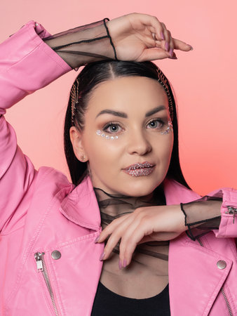 Portrait of a beautiful brunette in a leather jacket on a pink background. close-up. Bright make-up of glitter on the faceの写真素材