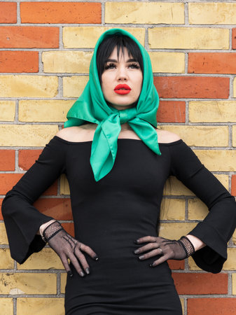 A woman with a perfect figure and red lips in a short black dress, a bright scarf on her head. Posing against the background of a brick wallの写真素材