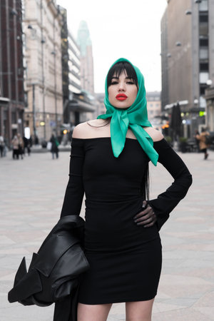 A woman with a perfect figure and red lips in a short black dress, a bright scarf on her head. Posing in the cityの写真素材