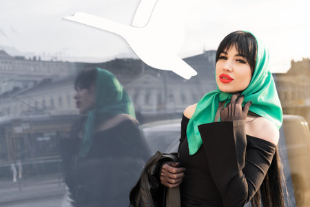 A woman with a perfect figure and red lips in a short black dress, a bright scarf on her head looks into the reflection of a shop windowの写真素材