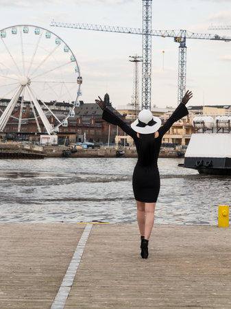 A woman with a perfect figure in a short black dress, on the pier in a white hat. Meets the ferry, waving his handsの写真素材