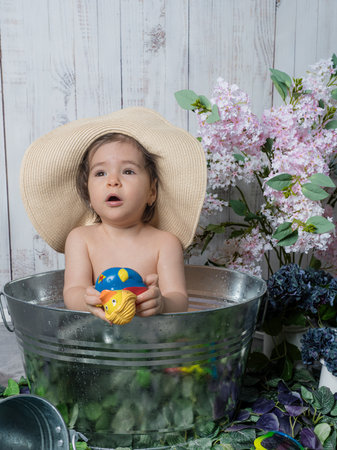 A little girl sits in a tub or basin, surrounded by rubber toys, wearing a large straw hat, creating a playful and charming scene.の写真素材