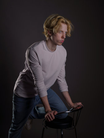 Studio portrait of a young man with blond hair, wearing a gray sweater and blue jeans, sitting on a chairの写真素材