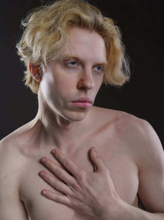 Artistic portrait of a young man with wavy blonde hair against a dark background. Bare shoulders, hand on chest. Thoughtful gaze, melancholic expression. Soft lighting highlights fの写真素材