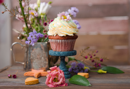 Cup cakes with cream cheese topping on beautiful wooden cake stand with flowers and wooden background behind it, spring mood still life photo for holiday designの写真素材